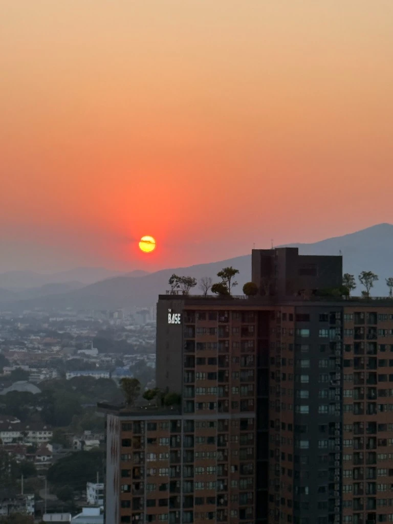 Burning-season haze at sunset over Chiang Mai, with the sun glowing through smog above mountain ridges and the city skyline.