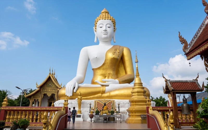 Large white seated Buddha with a gold robe on a temple terrace under a clear sky.