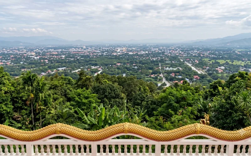 View over a forested hillside and city plain from a terrace with a white railing and golden naga handrail.