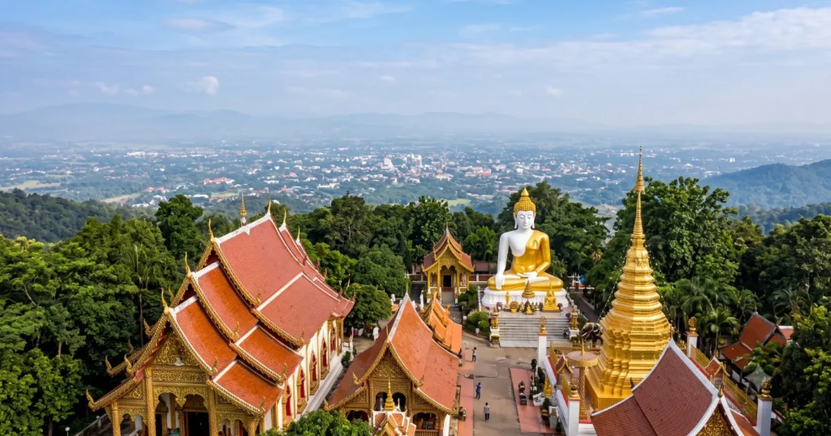 Elevated view of a hilltop temple complex with red-tiled roofs, a large white-and-gold seated Buddha and a golden chedi, overlooking a city valley.